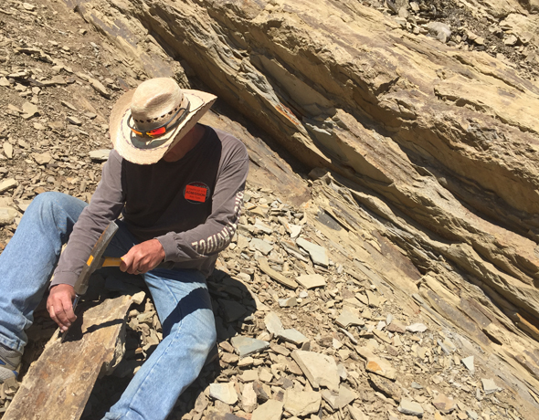 Man wearing cowboy-style hat breaking open rock in search of fossils.